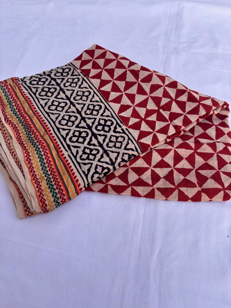 May include: A folded textile featuring a repeating red and cream triangular geometric pattern. The border displays a black floral motif on a cream background, finished with thin, parallel stripes in red, green, and yellow along the edge.