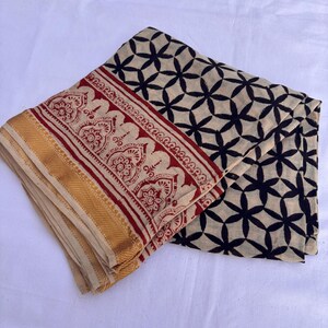 May include: A folded textile featuring a cream-colored base with a black geometric floral pattern. One edge displays a wide border with a red intricate botanical print and a solid gold-colored band along the finished hem of the fabric.