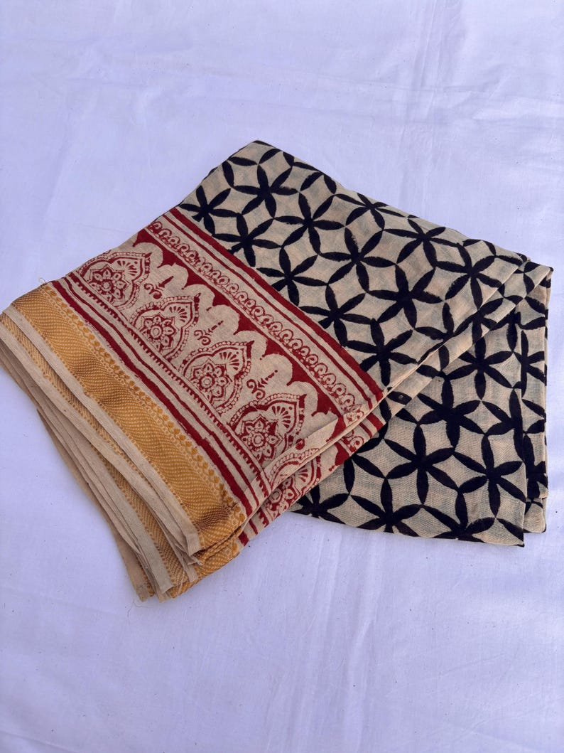 May include: A folded textile featuring a cream-colored base with a black geometric floral pattern. One edge displays a wide border with a red intricate botanical print and a solid gold-colored band along the finished hem of the fabric.