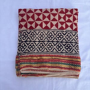 May include: A folded textile featuring a top section with a red and beige geometric triangle pattern, a middle band with a black floral motif on a beige background, and a bottom border with gold, green, and red horizontal stripes on beige fabric.
