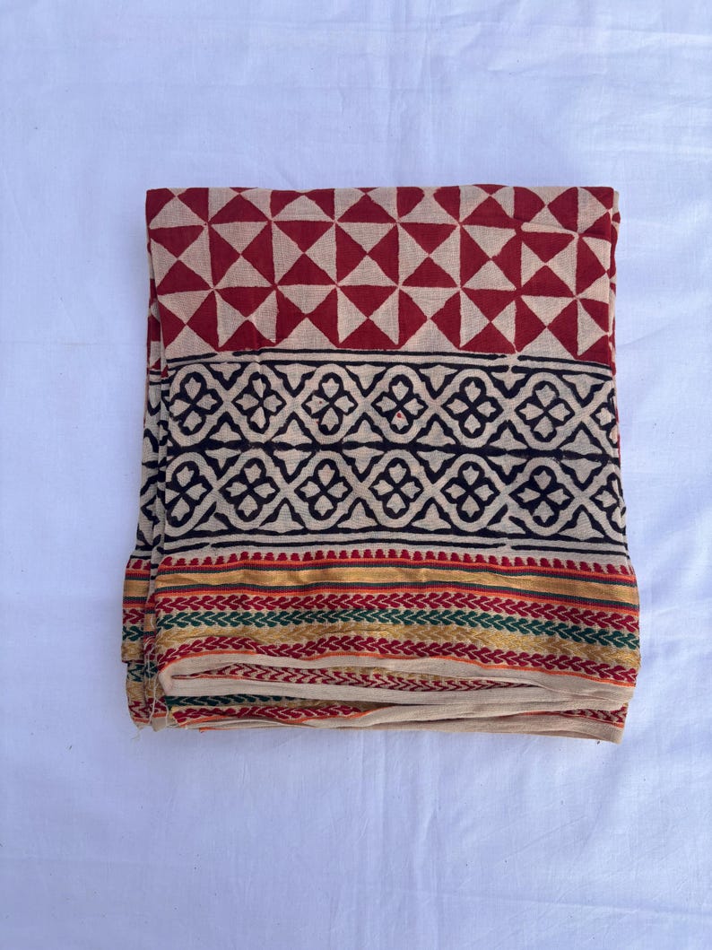 May include: A folded textile featuring a top section with a red and beige geometric triangle pattern, a middle band with a black floral motif on a beige background, and a bottom border with gold, green, and red horizontal stripes on beige fabric.