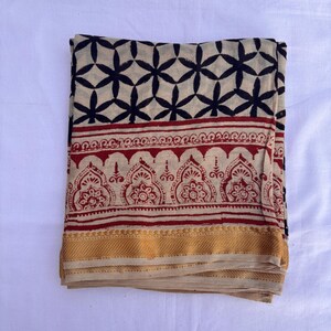 May include: A folded textile featuring a cream-colored base with a black geometric star pattern at the top, a central band of red intricate floral motifs, and a solid mustard yellow border along the bottom edge of the fabric piece.