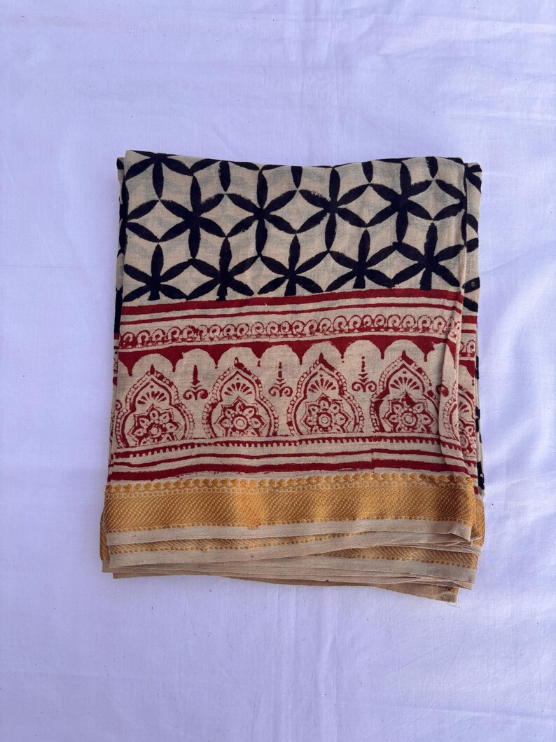 May include: A folded textile featuring a cream-colored base with a black geometric star pattern at the top, a central band of red intricate floral motifs, and a solid mustard yellow border along the bottom edge of the fabric piece.