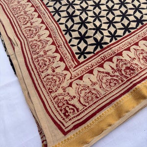 May include: A folded textile featuring a cream-colored base with a black geometric pattern of intersecting circles. The fabric is bordered by a wide red band with intricate floral motifs and finished with a textured mustard yellow edge trim.