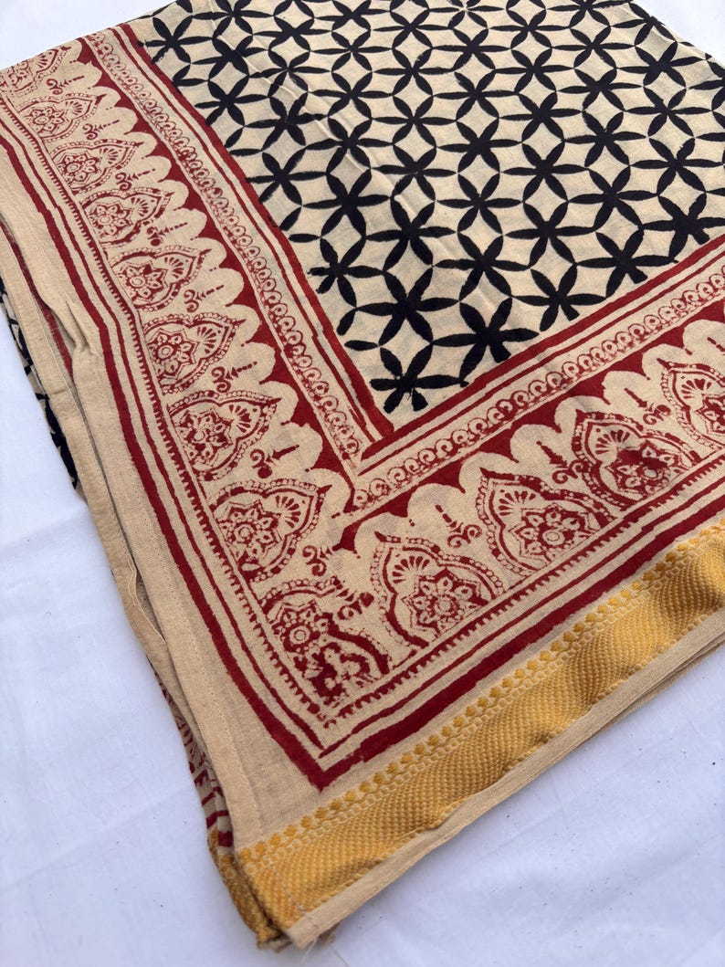May include: A folded textile featuring a cream-colored base with a black geometric pattern of intersecting circles. The fabric is bordered by a wide red band with intricate floral motifs and finished with a textured mustard yellow edge trim.