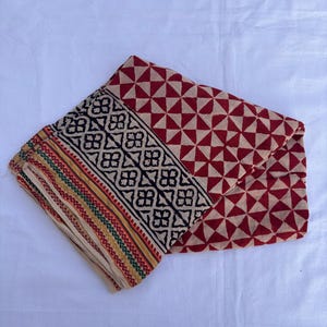 May include: A folded textile featuring a cream-colored base with a repeating red triangular geometric pattern. A wide black border displays a repeating floral motif, while the edge is finished with thin, parallel stripes in yellow, green, and red.