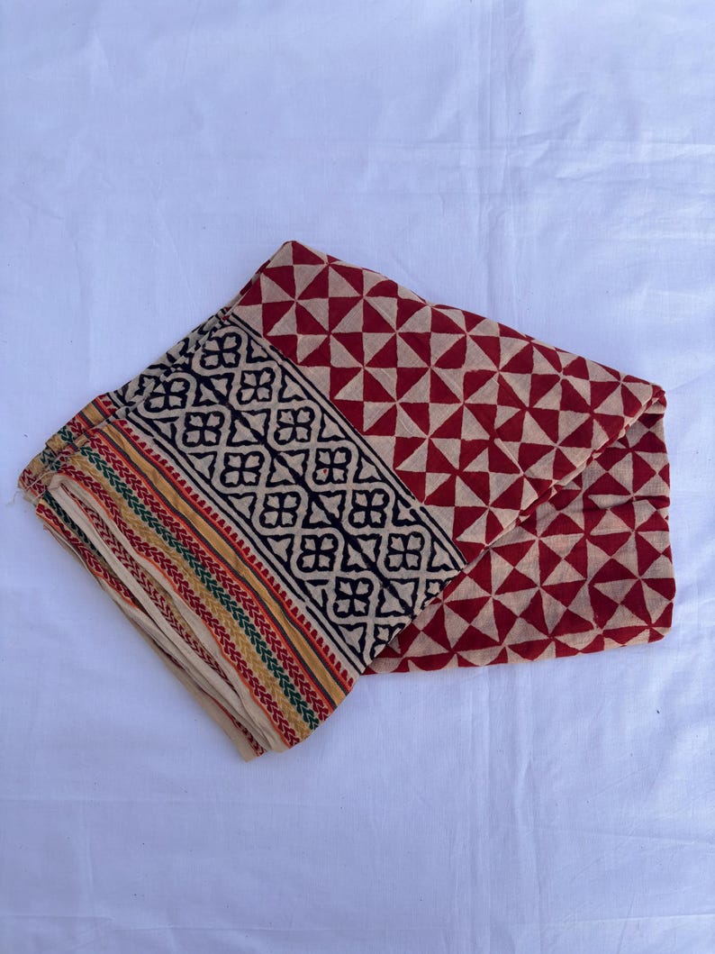 May include: A folded textile featuring a cream-colored base with a repeating red triangular geometric pattern. A wide black border displays a repeating floral motif, while the edge is finished with thin, parallel stripes in yellow, green, and red.