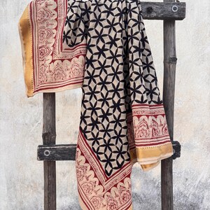 May include: A rectangular textile featuring a central pattern of black interlocking floral shapes on a cream background. The fabric is bordered by a red decorative print and finished with a solid gold-colored edge along the outer perimeter.