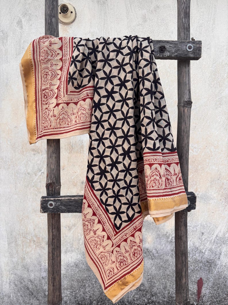 May include: A rectangular textile featuring a central pattern of black interlocking floral shapes on a cream background. The fabric is bordered by a red decorative print and finished with a solid gold-colored edge along the outer perimeter.