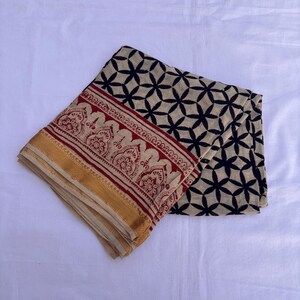 May include: A folded textile featuring a cream-colored base with a black geometric floral pattern. The border displays a red band with intricate decorative motifs and a solid mustard yellow edge, creating a distinct contrast in color and design.