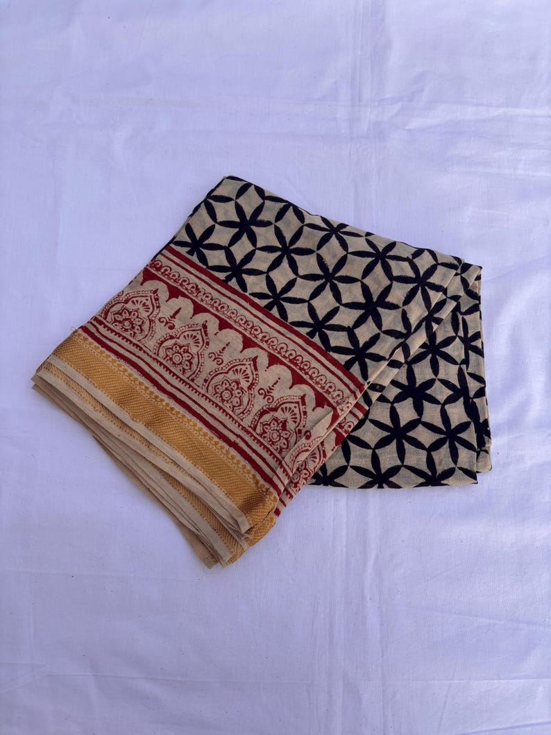May include: A folded textile featuring a cream-colored base with a black geometric floral pattern. The border displays a red band with intricate decorative motifs and a solid mustard yellow edge, creating a distinct contrast in color and design.