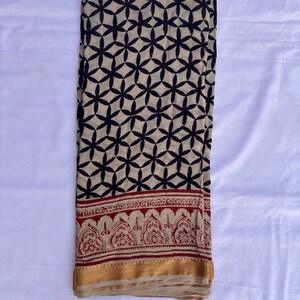 May include: A rectangular fabric textile featuring a beige base with a repeating black geometric floral pattern. The lower section displays a horizontal border with a red intricate botanical print, finished with a solid gold-colored hem at the edge.