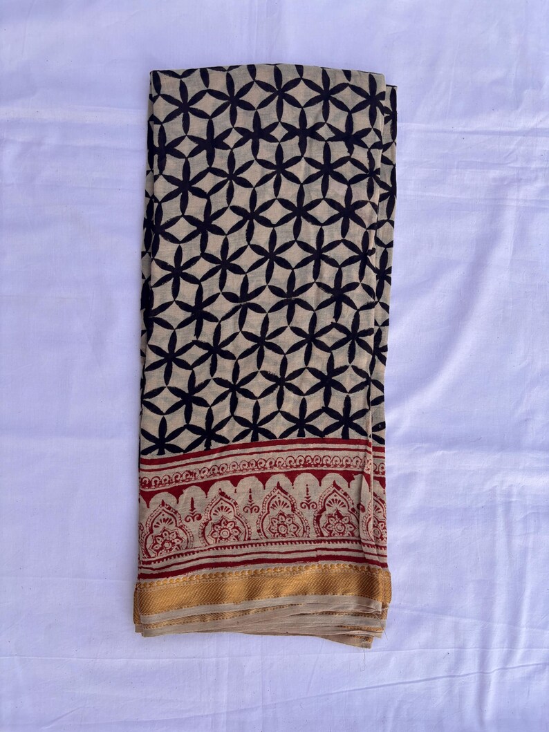 May include: A rectangular fabric textile featuring a beige base with a repeating black geometric floral pattern. The lower section displays a horizontal border with a red intricate botanical print, finished with a solid gold-colored hem at the edge.