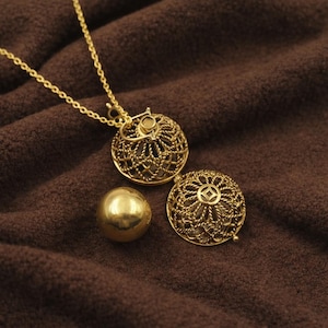Puede incluir: Un collar dorado con un colgante de diseño calado, junto con una esfera a juego y un segundo colgante. Las joyas se exhiben sobre un fondo de tela marrón, resaltando los intrincados detalles de las piezas.