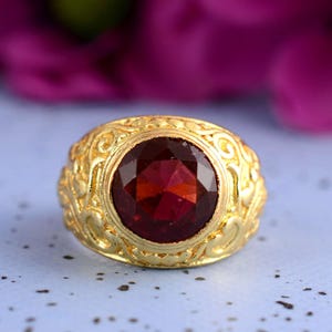 May include: A gold ring featuring a large, faceted, deep red gemstone. The ring has an ornate, engraved design around the band. The gemstone is the focal point, catching the light and creating a striking visual.
