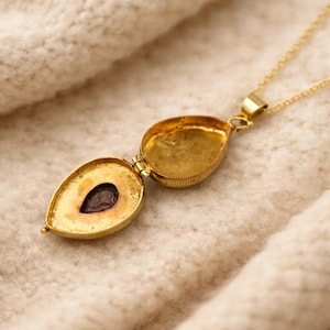 May include: A gold-colored locket necklace with an open, teardrop-shaped pendant. The pendant reveals a dark, oval-shaped stone inside. The necklace is on a cream-colored, textured surface.