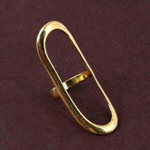 May include: A gold-colored ring with a long, open oval design. The ring has a band that wraps around the finger and a connecting bar within the oval. The ring is set against a dark maroon background.