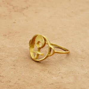 May include: A gold-colored ring featuring a stylized symbol. The ring's design incorporates a circular element with a cross-like shape and a curved line. The ring is set against a neutral background.