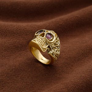 May include: A gold-colored skull ring with intricate detailing. The ring features purple-colored stones in the eye sockets. The skull design is the focal point, with detailed carvings on the sides. The ring is set against a brown background.
