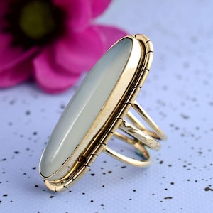 Op de afbeelding: Een goudkleurige ring met een grote, ovale, lichtblauwe edelsteen. De ring heeft een gedetailleerde, getextureerde band en een drievoudig bandontwerp. De edelsteen is gezet in een gouden bezel. De ring wordt getoond tegen een zachte blauwe achtergrond.