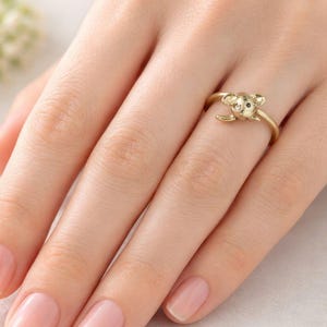 May include: A gold-colored ring featuring a koala bear design. The ring has a smooth band and the koala has small ears and a black eye. The ring is worn on a finger.