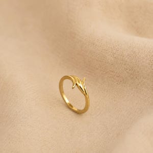 Puede incluir: Un anillo de tono dorado pulido con una banda delgada y abierta que se curva en un diseño delicado en forma de hoja o pétalo en la parte superior. La superficie metálica es lisa y reflectante, mostrando una forma continua y fluida.