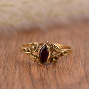 以下が含まれることがあります： 深紅色のマーキス型宝石をあしらったゴールドカラーのリング。宝石とバンドの周りには、花と葉のディテールが施されています。リングは木製の表面に置かれています。