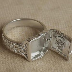May include: A silver-colored ring with an ornate band and a rectangular compartment that opens to reveal a skull design. The ring has a detailed, engraved pattern on the band.