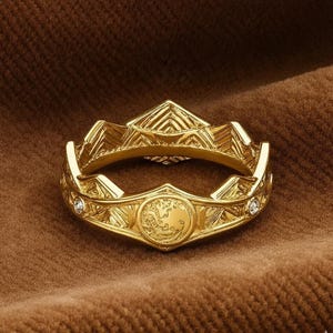 May include: A gold ring with a crown design. The ring has detailed geometric patterns and small, clear gemstones. The ring is set against a brown background, highlighting its details and craftsmanship.