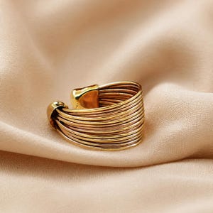 Könnte beinhalten: Ein goldfarbener Ring mit einem einzigartigen Design. Der Ring hat mehrere parallele Bänder, die sich um den Finger wickeln und ein strukturiertes Aussehen erzeugen. Der Ring liegt auf einem weichen, neutralfarbenen Stoff.