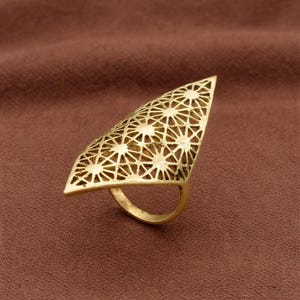 Puede incluir: Un anillo dorado con una gran cara triangular con un diseño geométrico calado. La banda del anillo tiene una forma simple y redondeada, y la cara está adornada con patrones en forma de estrella. El anillo está sobre un fondo marrón.