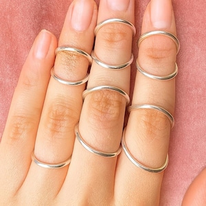 May include: A hand with several silver rings. The rings are thin, curved bands that encircle the fingers, offering a minimalist aesthetic. The rings are worn on multiple fingers, highlighting their design and shape.
