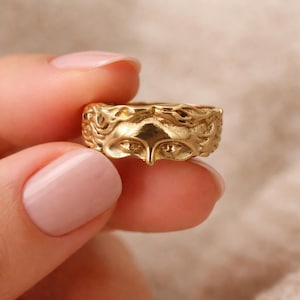 Op de afbeelding: Een goudkleurige ring met een gedetailleerd gezichtsontwerp. De ring heeft een textuuroppervlak met gelaatstrekken, waaronder ogen en een neus. De ring wordt tussen de vingers gehouden en toont zijn ingewikkelde vakmanschap en ontwerp.