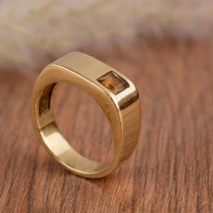Può includere: Anello color oro con una gemma rettangolare color ambra. L'anello ha una finitura liscia e lucida e un design classico e minimalista. L'anello è mostrato su una superficie di legno.