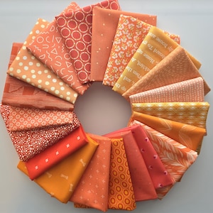 May include: A circular arrangement of folded fabric squares in various shades of orange, peach, and coral. The fabrics feature diverse patterns, including polka dots, geometric shapes, and text. The arrangement is set against a white background, creating a visually appealing display.