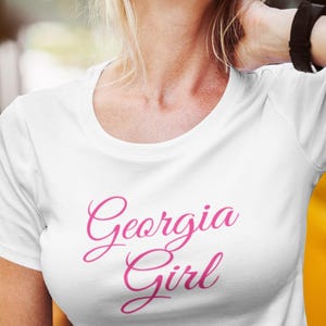 May include: White t-shirt with the cursive text "Georgia Girl" in pink. The shirt has a crew neck and short sleeves. The image shows the shirt on a person.