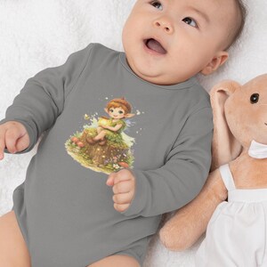 Op de afbeelding: Een grijze babybody met lange mouwen met een kleurrijke illustratie van een fee die op een boomstronk zit. De fee draagt een groene jurk en houdt een gouden voorwerp vast. Een pluchen konijnenspeelgoed staat naast de baby.