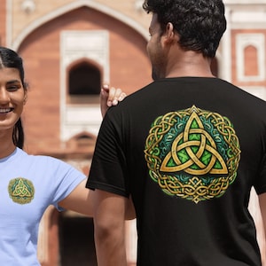 Könnte beinhalten: Schwarzes T-Shirt mit einem gold-grünen keltischen Knotenmuster auf dem Rücken. Ein hellblaues T-Shirt mit einem kleineren, passenden Design ist ebenfalls zu sehen. Die Shirts werden von zwei Personen getragen, mit einem verschwommenen Hintergrund.