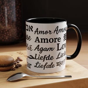 May include: White ceramic mug with black interior and handle. The mug features the word "Love" in multiple languages. A silver spoon and a macaron are on a wooden surface next to the mug.