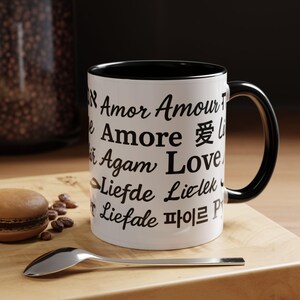 May include: White ceramic mug with black interior and handle. The mug features the word "Love" in multiple languages in black font. A silver spoon and a macaron are on a wooden surface.