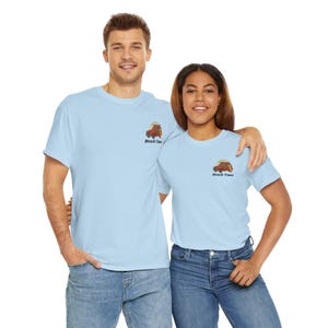 May include: Pale blue t-shirts featuring a small graphic of a vintage van with a surfboard, and the words "Beach Time". The shirts are worn with blue jeans. The image shows two people.