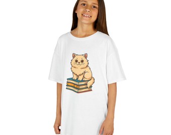 May include: White t-shirt featuring a cartoon cat sitting on a stack of books. The cat is light brown with a fluffy appearance and large eyes. The books are in shades of orange, blue, and yellow. The t-shirt is a casual style.