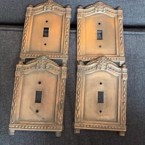 May include: Four antique bronze-colored light switch covers. Each cover has a rectangular opening for the switch and is framed with ornate detailing. The top of each cover features a decorative crest. The covers are arranged on a gray surface.