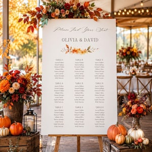 Pode incluir: Um cartaz branco de disposição de assentos de casamento num cavalete de madeira com o texto Please Find Your Seat, OLIVIA & DAVID, e uma lista de nove mesas com nomes. O cartaz é decorado com um motivo de folhas de outono e flores.