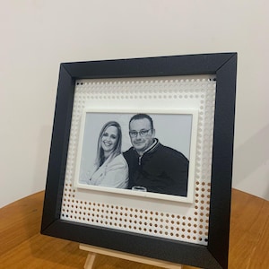 Pode incluir: Uma moldura preta com uma foto a preto e branco de um casal, apresentada num pequeno cavalete de madeira. A moldura tem uma borda branca perfurada.