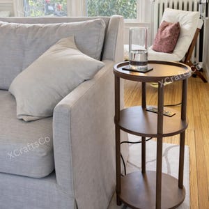 May include: A three-tiered, dark brown side table with a round top and shelves. A clear glass with water sits on the top shelf. The table is next to a light gray sofa and a white chair with a pink pillow.