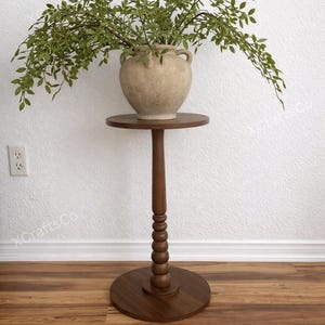 May include: A decorative, dark brown wooden plant stand with a round top and base, connected by a turned, beaded column. A beige, textured vase with green foliage sits on top. The stand is approximately 30 inches tall.