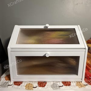 May include: White wooden bread box with two glass doors with a ribbed texture. The box has a knob on each door. The text "XCraftsCo" is printed on the box.