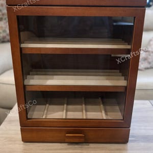 May include: A wooden display case with a glass front and multiple shelves. The case is a rich brown color and features a drawer at the bottom. The text "XCraftsCo" is visible on the case. The shelves appear to be lined with a light-colored material.