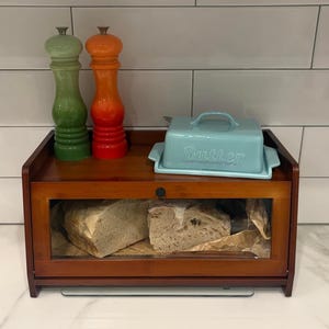 May include: A wooden bread box with a glass front, holding two loaves of bread. A light blue butter dish and two colorful salt and pepper grinders sit on top. The grinders are green and orange.